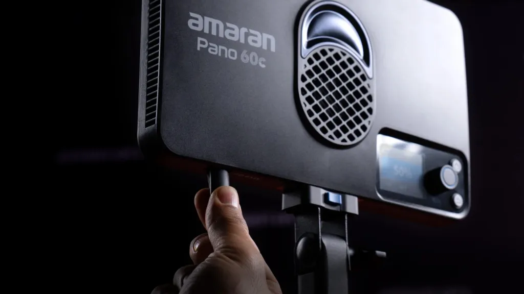 Крупний план чорної LED-панелі amaran Pano 60c з логотипом бренду, параболічним рефлектором і вентиляційною решіткою, на екрані відображається рівень яскравості 50 відсотків, палець оператора торкається кнопки живлення
