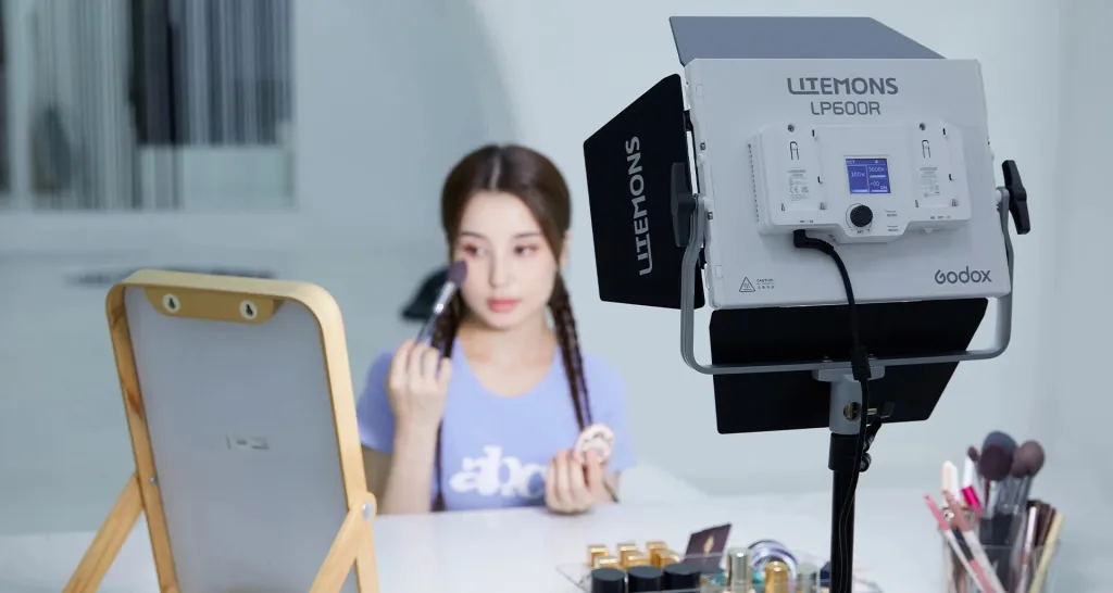LED-панель Godox Litemons LP600R на стійці, дівчина на задньому плані наносить макіяж перед дзеркалом, на екрані приладу відображено 5600K, 100 відсотків потужності та підключений кабель живлення